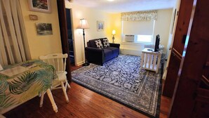 A cozy living room with a wooden floor, a blue sofa, a white chair, a dining table with a leaf-patterned tablecloth, and a patterned rug. - Beautiful Historic Cottage, Hideaway (Mount Dora)