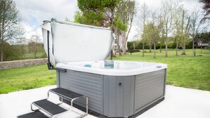 Un jacuzzi con escalón y tapa, en un patio de concreto con césped y árboles al fondo.
