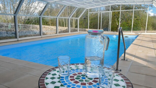 Zona de piscina con cerramiento de cristal, mesa con baldosas, jarra de vidrio y vasos, y escalera metálica.