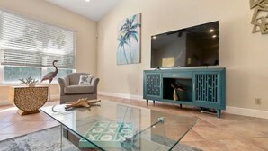 A living room with a glass coffee table, a teal cabinet, a flat-screen TV, a sofa, and a window with blinds. - Singer Island Beach Oasis: Heated Pool, Privacy Fence & Open Concept (West Palm Beach)