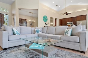 A modern living room with a sectional sofa, a glass coffee table, and a kitchen in the background. - Singer Island Beach Oasis: Heated Pool, Privacy Fence & Open Concept (West Palm Beach)
