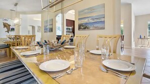 A dining area with a long table set for four, featuring a modern chandelier, wooden chairs, and a painting on the wall. - Singer Island Beach Oasis: Heated Pool, Privacy Fence & Open Concept (West Palm Beach)