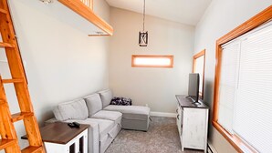 Une pièce de style loft avec un plafond haut, un ventilateur de plafond, une petite échelle en bois, un canapé d’angle, une armoire blanche et une grande fenêtre avec des stores.