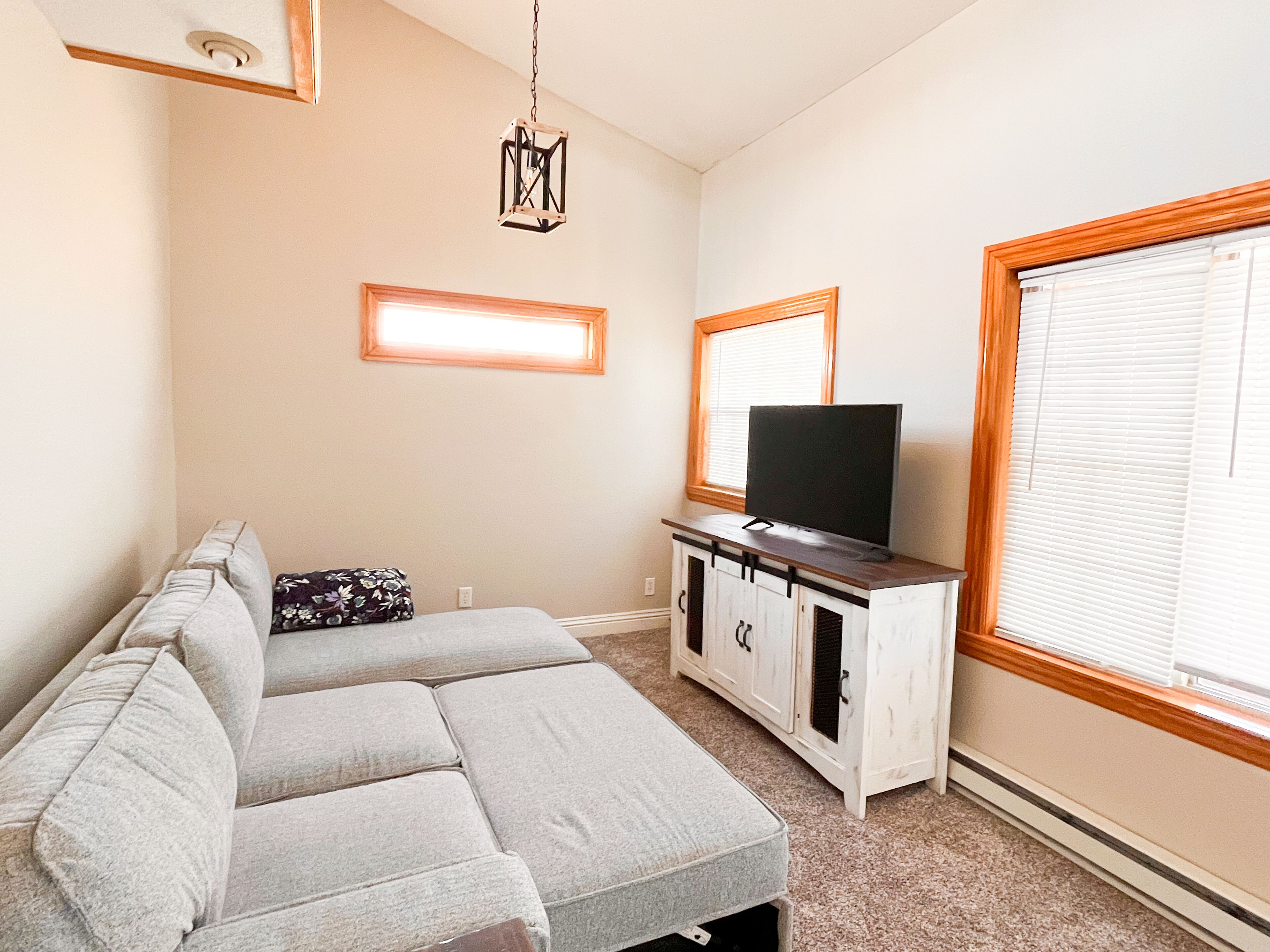 Un salon comprenant un canapé d’angle, un téléviseur à écran plat posé sur un meuble en bois, un luminaire suspendu et une grande fenêtre avec des stores.