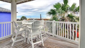 Un espace extérieur abrité, doté d’une terrasse en bois, de chaises Adirondack blanches et offrant une vue sur des palmiers et un bâtiment bleu.