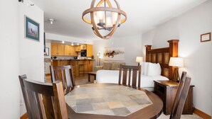 A dining area with a round table, wooden chairs, and a chandelier. There is a kitchen in the background with wooden cabinets and a microwave.