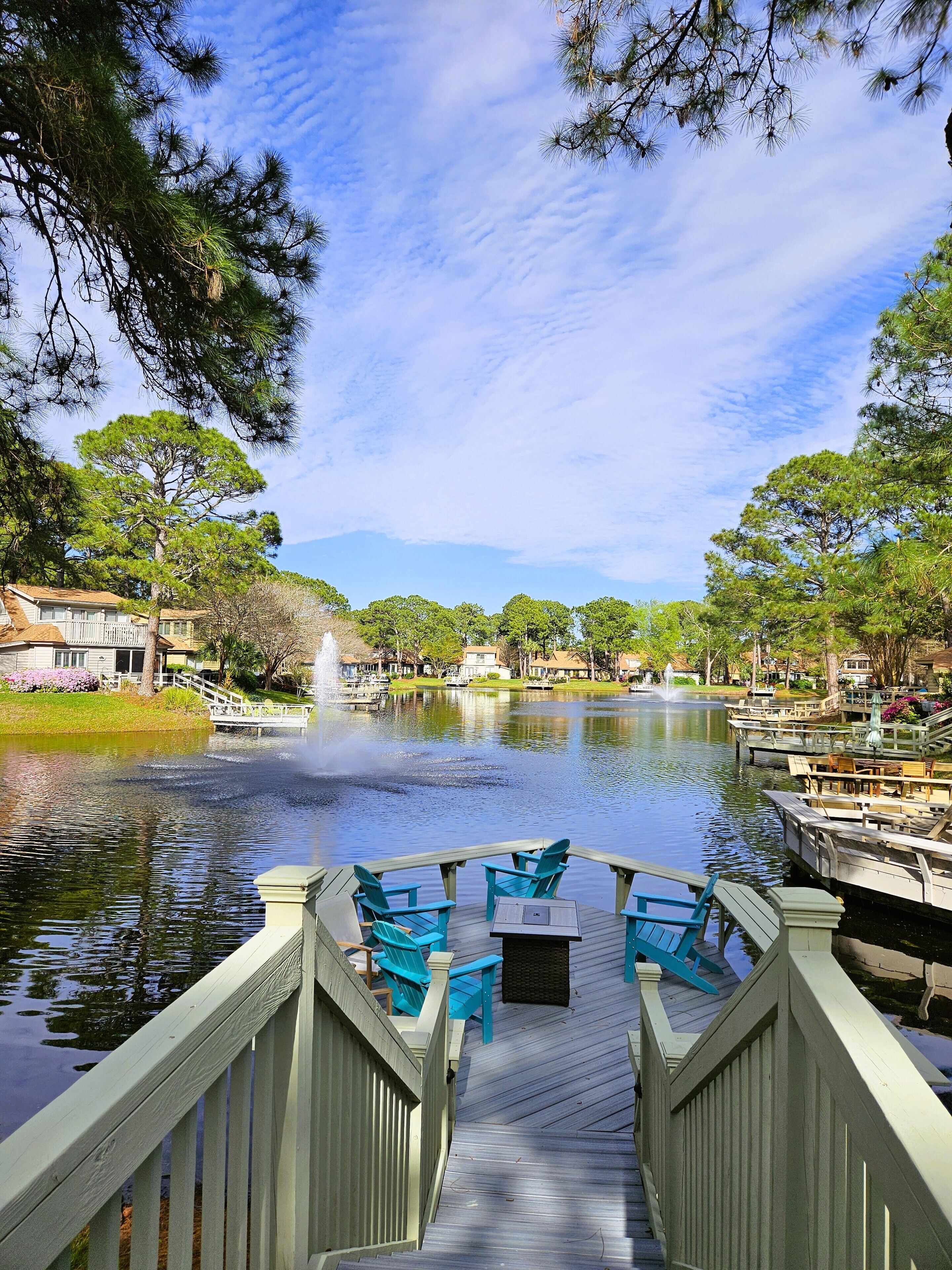 A wooden deck with chairs and tables overlooking a calm lake, surrounded by trees and a clear sky.
