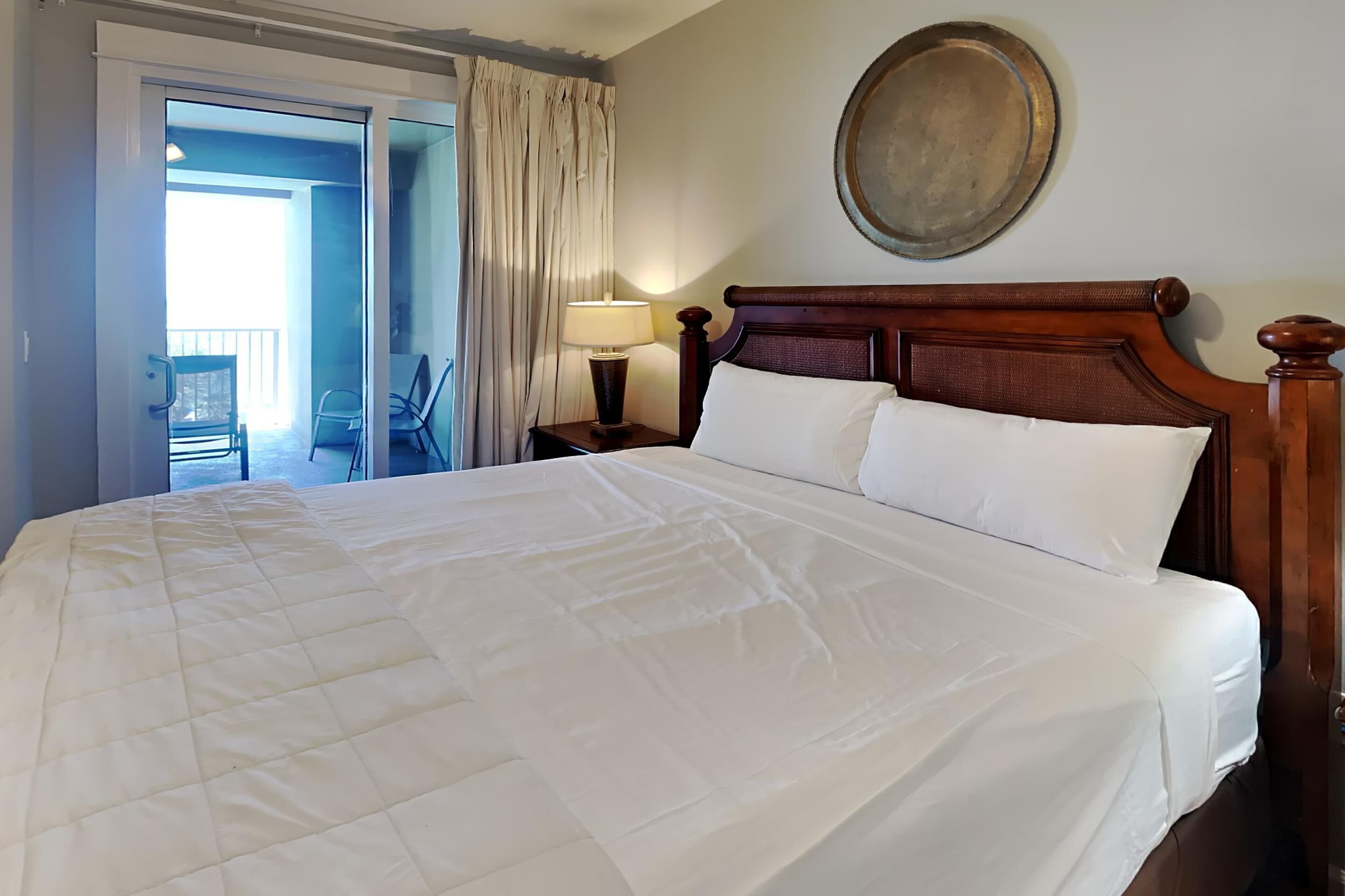 A neatly made bed with a wooden headboard, a bedside lamp, and a view of a balcony through an open door.