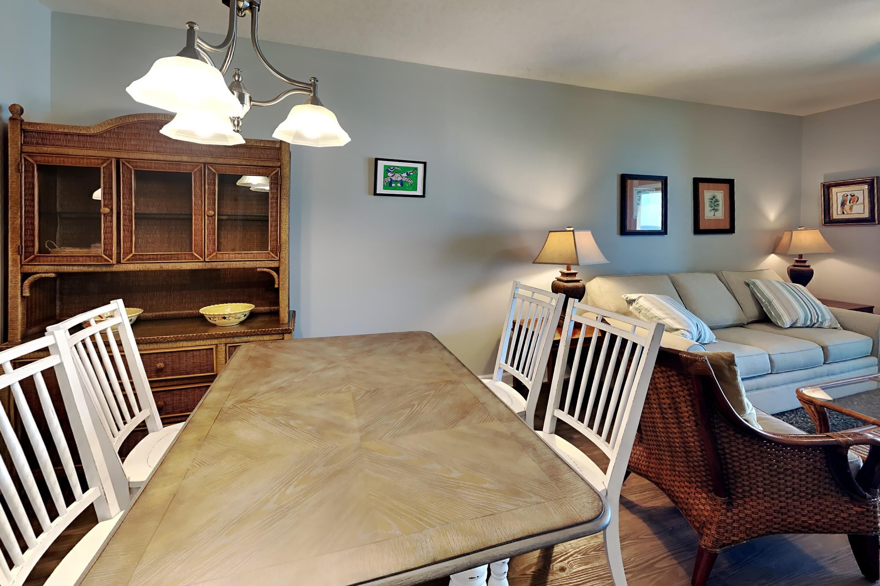 A dining area with a wooden table and white chairs, a wicker sofa, and a cabinet with glass doors.