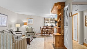 Una sala de estar con sofá, sillón y mesa de comedor con sillas. Hay un televisor, una lámpara y un cuadro en la pared.