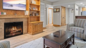 Una sala de estar con chimenea, un televisor montado en una pared de madera, un sofá y una mesa de centro de vidrio.