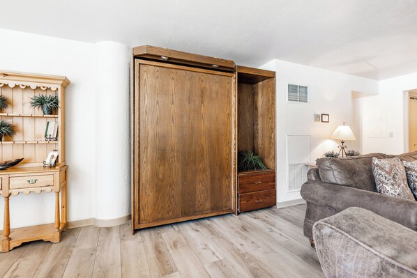 A room with a wooden wardrobe, a wooden side table with plants, a grey sofa, and a lamp.