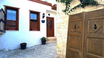 Un cortile con pavimento in pietra, un edificio bianco con persiane di legno e un muro in pietra con una porta di legno.