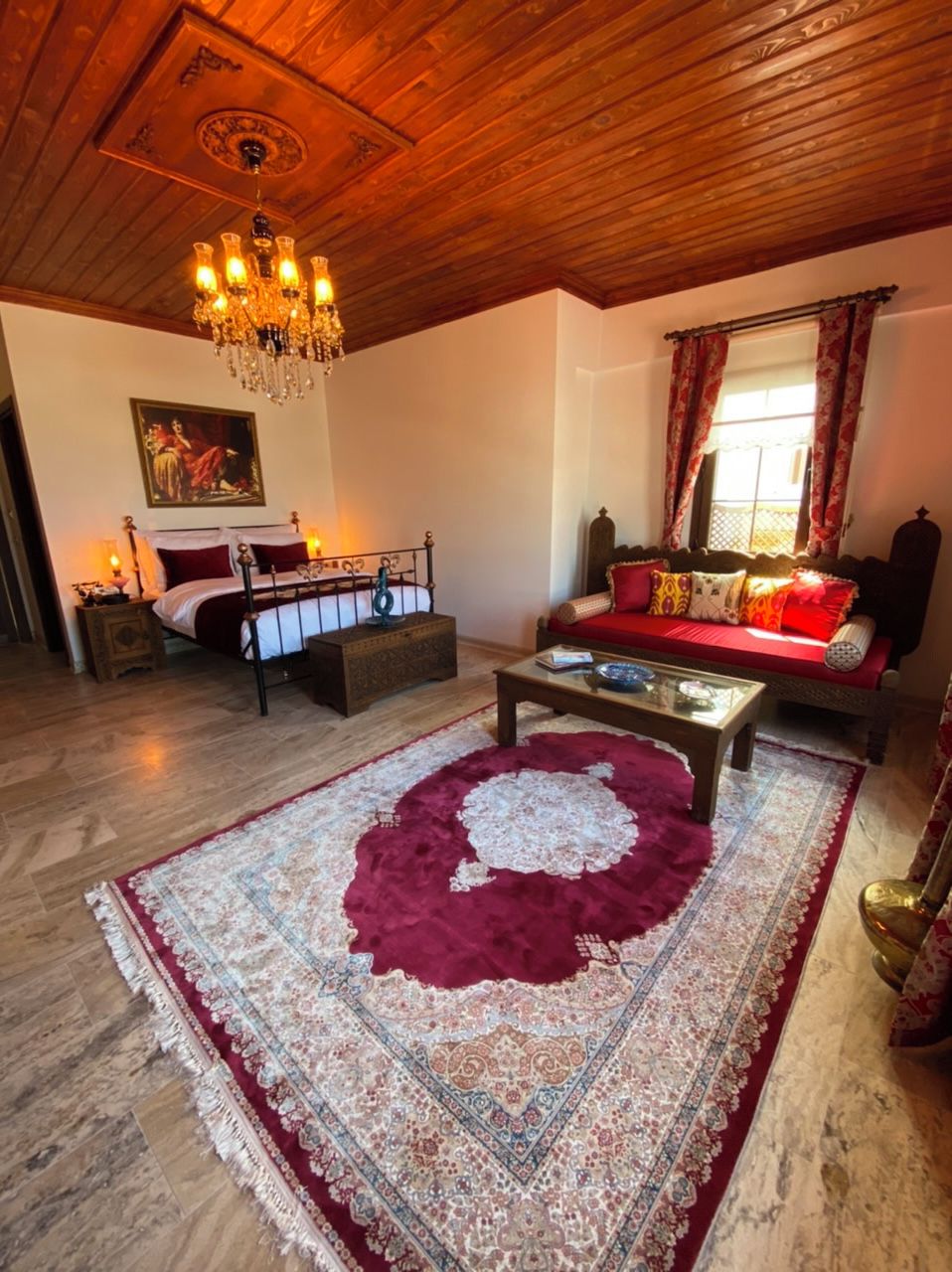 Une pièce avec un plafond en bois, un lustre, un lit recouvert d’une courtepointe rouge et blanche, un canapé avec des coussins rouges, une table basse et un tapis à motifs.