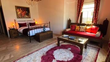 Une pièce avec un plafond en bois, un lustre, un lit recouvert d’une courtepointe rouge et blanche, un canapé avec des coussins rouges, une table basse et un tapis à motifs.