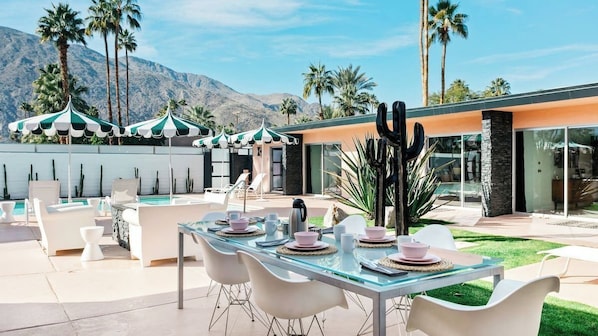 A poolside dining area with a glass table, white chairs, and green and white striped umbrellas.