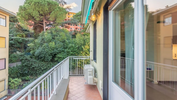A balcony with a view of a lush garden and buildings in the background.