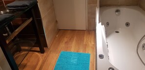 A bathroom with a wooden floor, a blue bath mat, a white bathtub, and a shelf with folded towels.