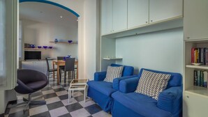 A living room with a checkered floor, a blue sofa, a small table, and a chair. There is a kitchen area in the background with a dining table and chairs.