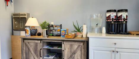 A hotel reception area with a coffee station, a refrigerator, a lamp, a plant, and a magazine rack.