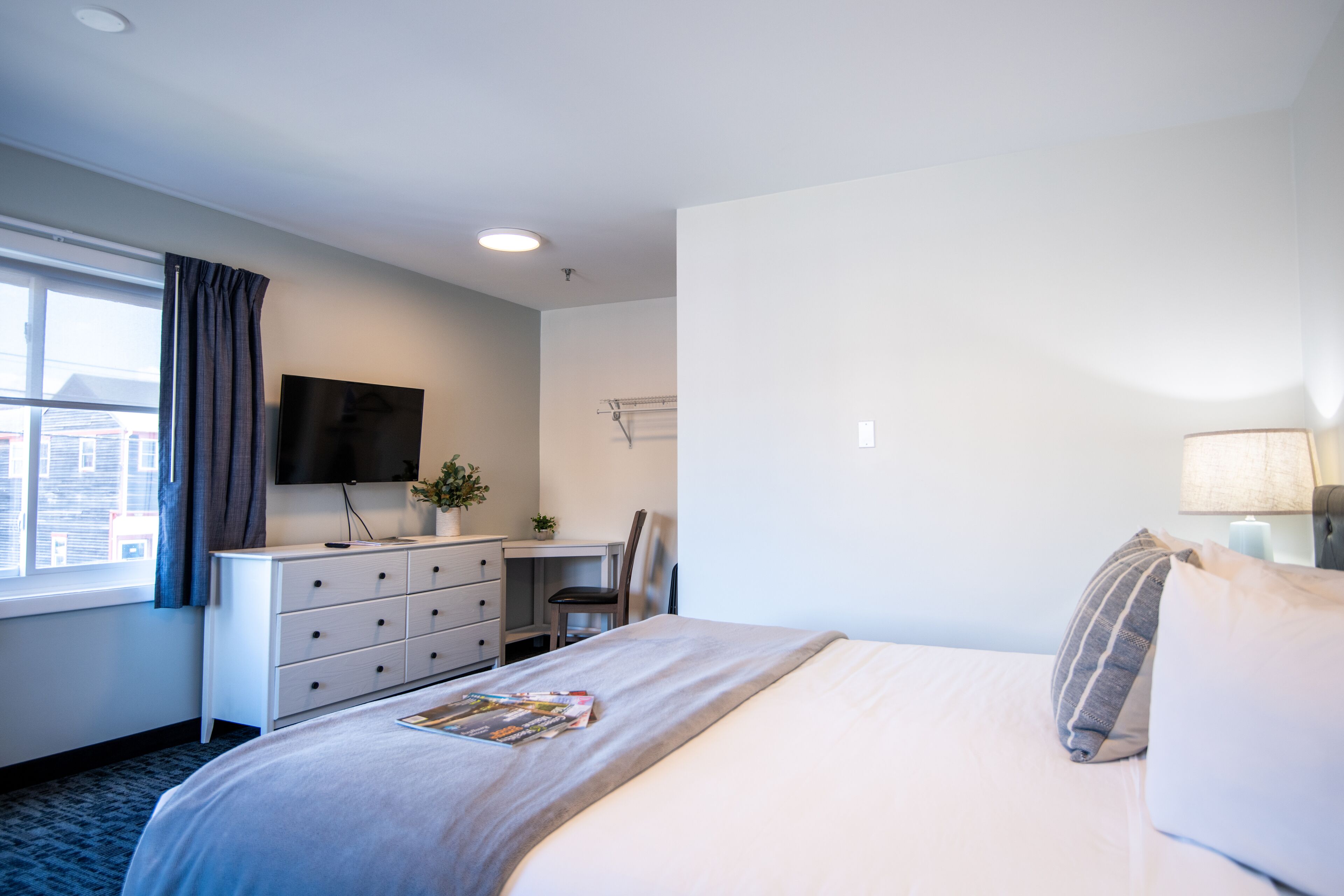 Quarto de hotel com cama, escrivaninha, cadeira, televisão e janela com cortinas.