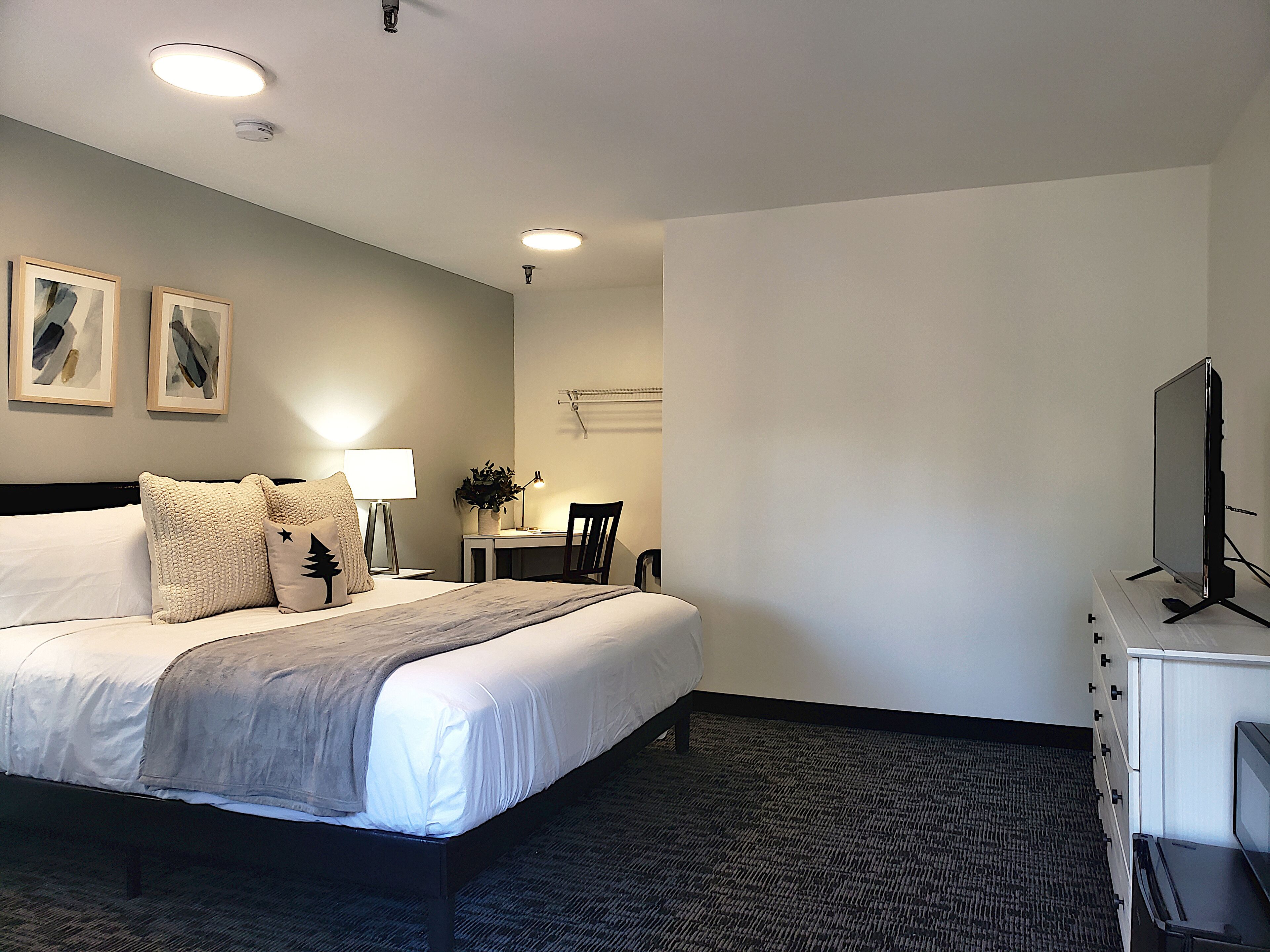 A hotel room with a bed, desk, chair, TV, and artwork on the wall.