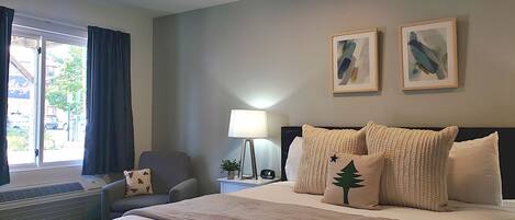 A hotel room with a bed, a chair, a desk with a lamp, and two framed pictures on the wall.
