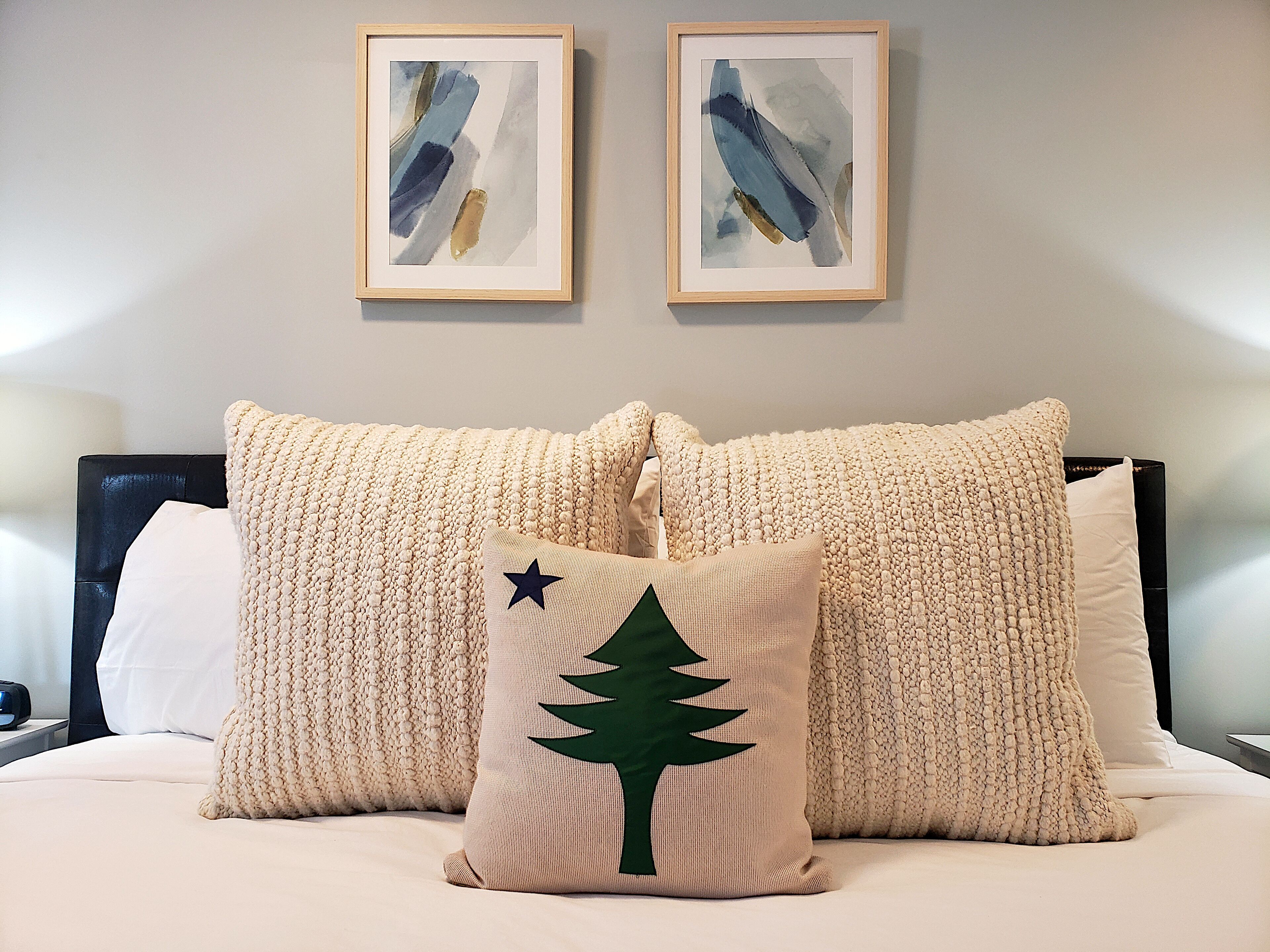 A neatly made bed with two large, textured pillows and a smaller pillow with a tree design. There are two framed artworks above the bed.