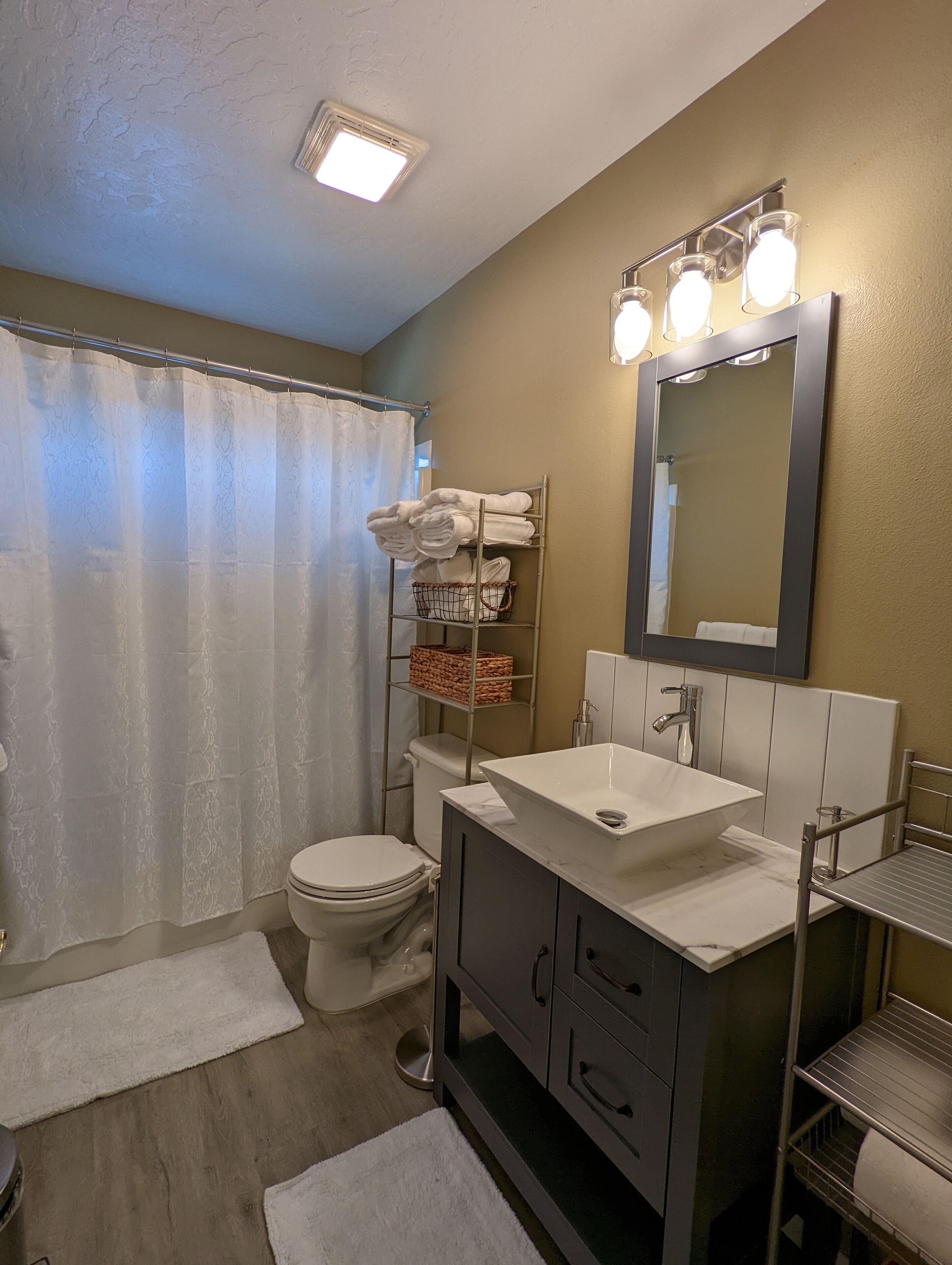 A bathroom with a toilet, sink, and shower curtain. There is a towel rack with towels and a basket.