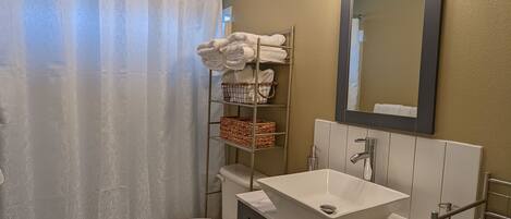 A bathroom with a toilet, sink, and shower curtain. There is a towel rack with towels and a basket.