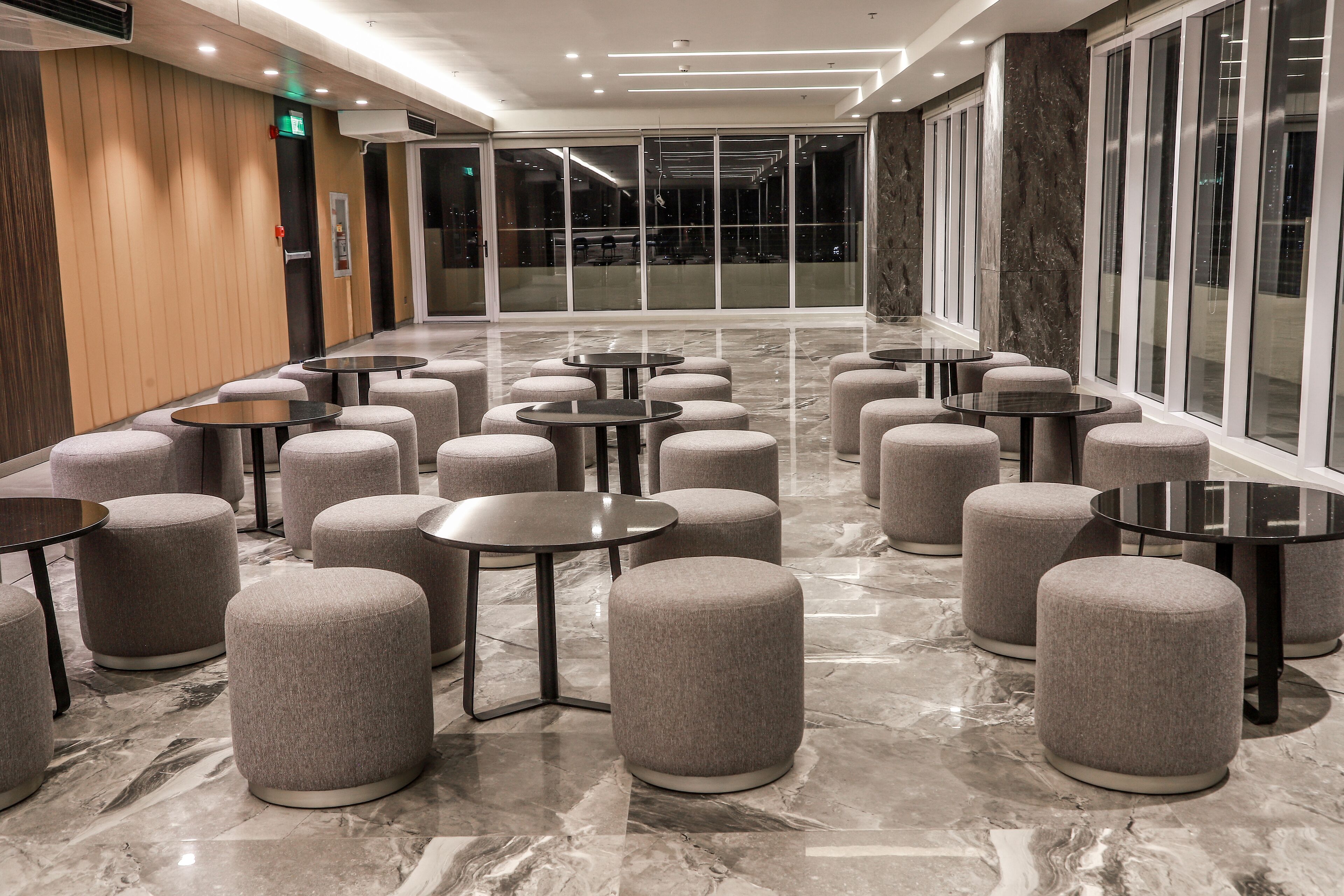 A modern hotel lobby with round tables and ottomans arranged in rows, large windows, and a marble floor.