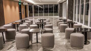 A modern hotel lobby with round tables and ottomans arranged in rows, large windows, and a marble floor.