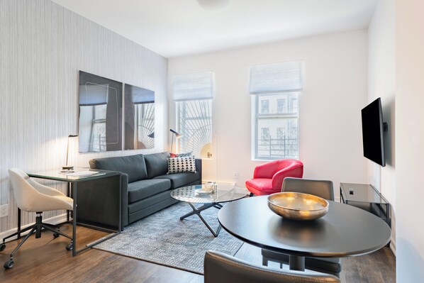 A modern living room with a sofa, coffee table, and red chair. There is a flat-screen TV mounted on the wall and a desk with a lamp.