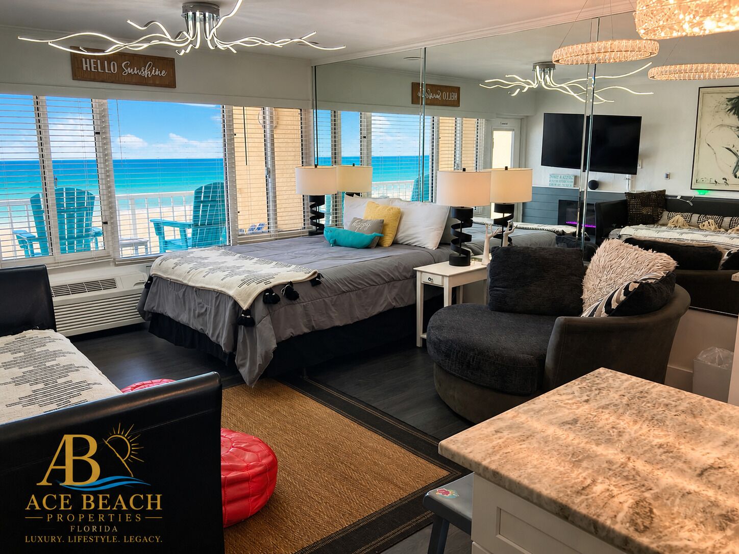 A hotel room with a bed, a desk, a chair, a red ottoman, and a view of the ocean.
