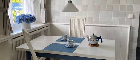A dining area with a white table, blue placemats, two white chairs, a hanging pendant light, and a window with curtains.