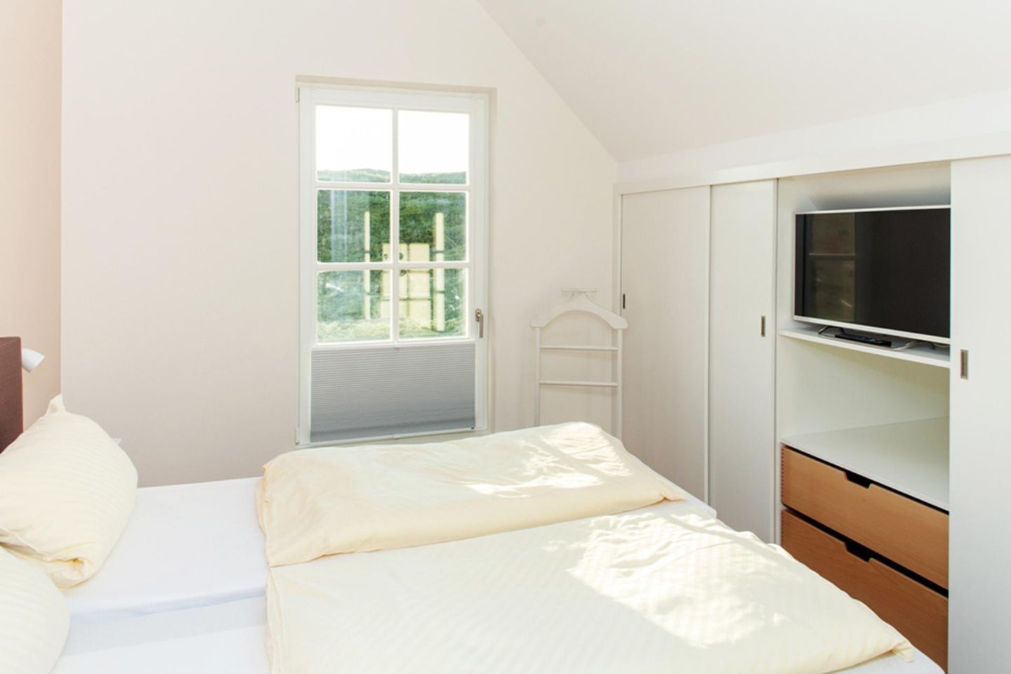 A bedroom with a bed, a TV, a wardrobe, and a window with a view of greenery.