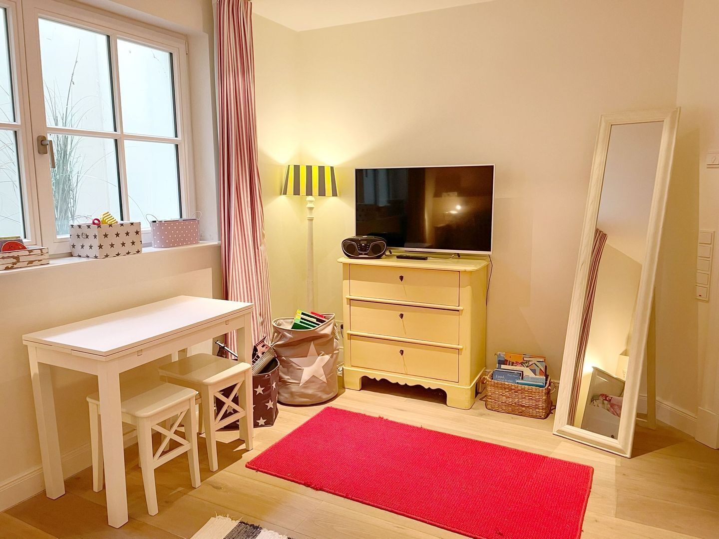 Una habitación con un escritorio blanco, televisión, espejo y una alfombra roja.
