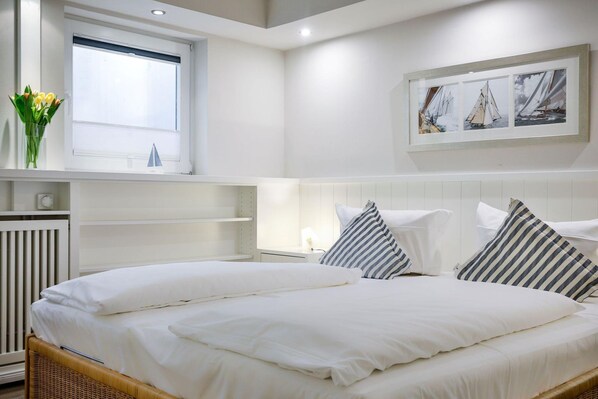 A neatly made bed with white linens, a window with blinds, a wall-mounted painting of sailboats, and a bedside table with a lamp. - Raantem Hüs App. 05 - Raantem Hüs (Sylt)