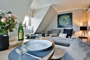 A dining area with a table set for two, a bottle of wine, and a vase of flowers. A grey sofa and a painting are in the background. - House Heefkant App. 08 - Heefkant (Sylt)