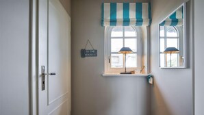 A hallway with a door, a window with a striped valance, a small shelf with a lamp, and a sign that says 'TO THE BEACH'.
