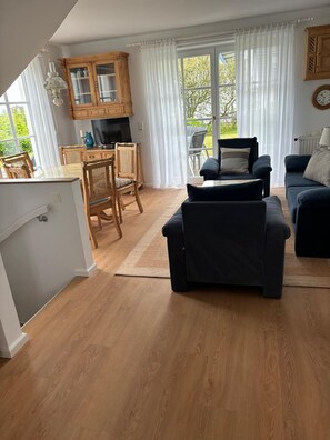 Une cuisine moderne avec des meubles en bois, un coin repas doté d’une table en verre et un salon avec un canapé foncé et un téléviseur.