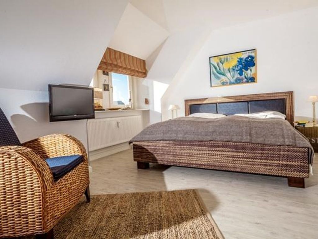 A bedroom with a large bed, a television, a wicker chair, and a painting on the wall.