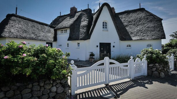 A traditional white house with a steeply pitched thatched roof, a white picket fence, and a stone wall in front. - House Lore, Germany (Sylt)