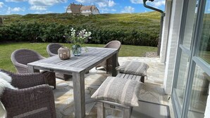 Een terras buiten met een houten tafel en rieten stoelen, een vaas met bloemen, en een uitzicht op een huis op de achtergrond.