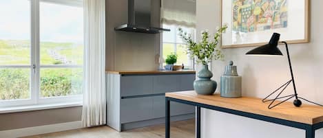 A modern kitchen with a large window, grey cabinets, a black range hood, and a table with a lamp and vases.