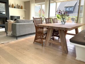 Ein modernes Wohnzimmer mit grauem Sofa, Esstisch aus Holz, Korbstühlen und Blick auf ein Haus durch große Fenster.
