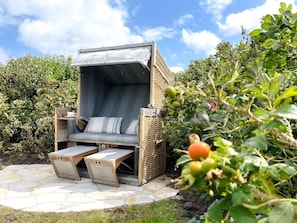 A cozy outdoor seating area with a cushioned bench and a small table, surrounded by lush greenery and a clear sky. - Beach House - Beach House AS/20 (Sylt)