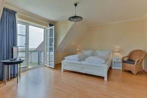 Une chambre avec un lit blanc, une chaise en osier, une petite table de chevet et une vue sur l’extérieur grâce à une grande fenêtre.