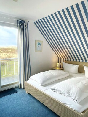 A hotel room with a bed, a nightstand, a lamp, a framed picture, and a view of the outdoors. - Im Dünengrund App. 14 and 15 - Dünengrund (Sylt)