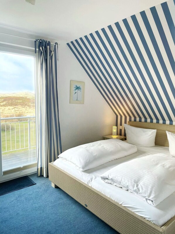 A hotel room with a bed, a nightstand, a lamp, a framed picture, and a view of the outdoors. - Im Dünengrund App. 14 and 15 - Dünengrund (Sylt)